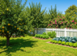 A lush, verdant garden, brimming with a bountiful array of fruit trees laden with ripe fruit, vibrant vegetable patches, and a profusion of colorful flowering plants, is beautifully framed by a pristine white picket fence beneath a clear, sunny blue sky